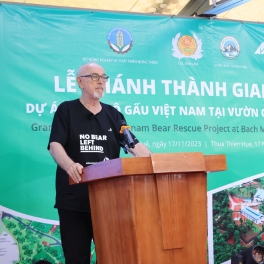 Person in a "No Bear Left Behind" t-shirt speaking at a podium during a bear rescue project event with a green banner.