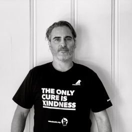 Person wearing a black t-shirt with "The Only Cure Is Kindness" text, standing against a white wall.