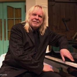 Person with long blonde hair in a pinstripe suit sitting beside a grand piano in a warmly lit room.