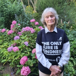 Person in a "The Only Cure Is Kindness" t-shirt standing in a garden with blooming pink hydrangea flowers.