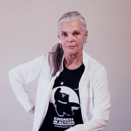 Person in a white jacket and black "Kindness in Action" t-shirt sitting against a plain wall.