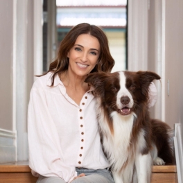 Person in a white buttoned shirt sitting on wooden stairs beside a brown and white dog indoors.