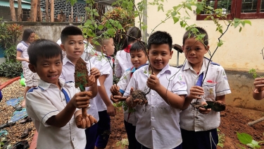 School children in uniforms hold young plants and roots, participating in an outdoor tree-planting activity.