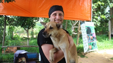Person holds a tan dog in their arms under an orange canopy at an outdoor rescue or adoption event.
