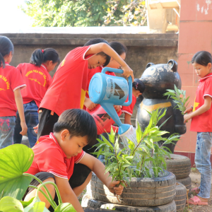 A group of young Vietnamese school children in red tshirts work busily in a garden, in the foreground, one is watering plants and another is crouched down and planting herbs.