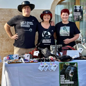 Three people standing behind a table with bear rescue merchandise and awareness posters at an outdoor event.