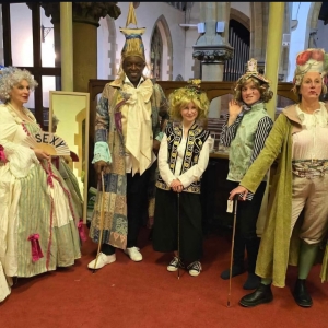Group dressed in elaborate historical costumes posing inside a church with red carpeting and stone columns.