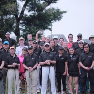Group of people in black shirts posing together outdoors on a golf course, holding golf clubs.