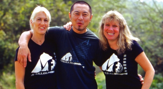 Three people wearing Animals Asia shirts stand together outdoors with arms around each other.