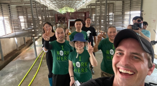 Group of people wearing green "I Work Here" shirts posing inside an animal shelter with metal enclosures.