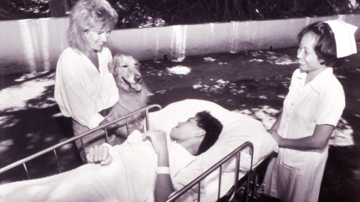 Golden retriever and two women stand beside a hospital bed outdoors, providing comfort to a patient lying under a blanket
