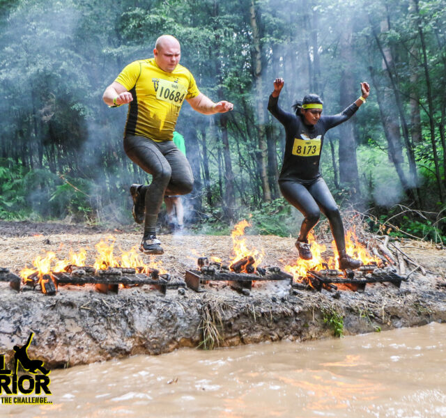 Two runners leap over flaming logs during a muddy outdoor obstacle race in a forest setting.