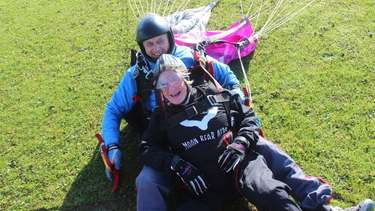 Older woman with moon bear hero sweater lying on grass with man behind her with a parachute, both are laughing.