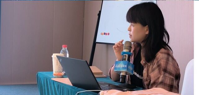 Woman presenting, seated at blue covered table with laptop and microphone, whiteboard in background.