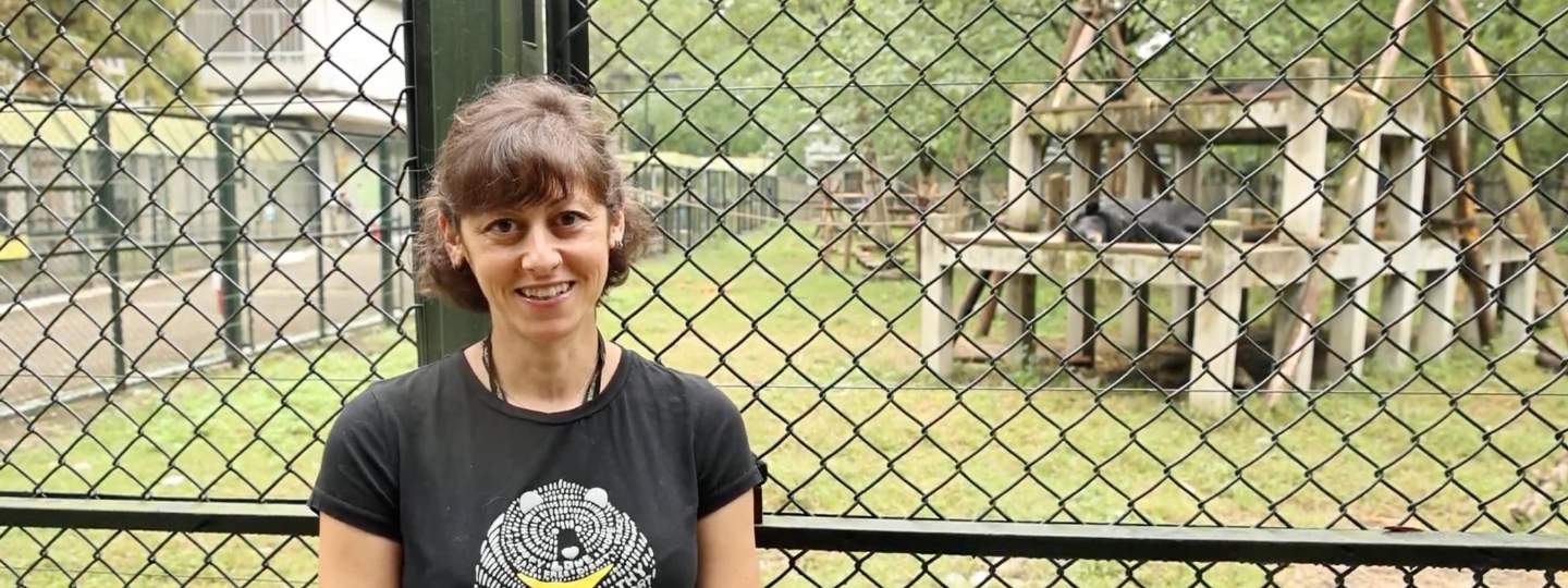 A caucasian woman with short brown hair stands smiling in front of a grassy bear enclosure as a bear relaxes in background.