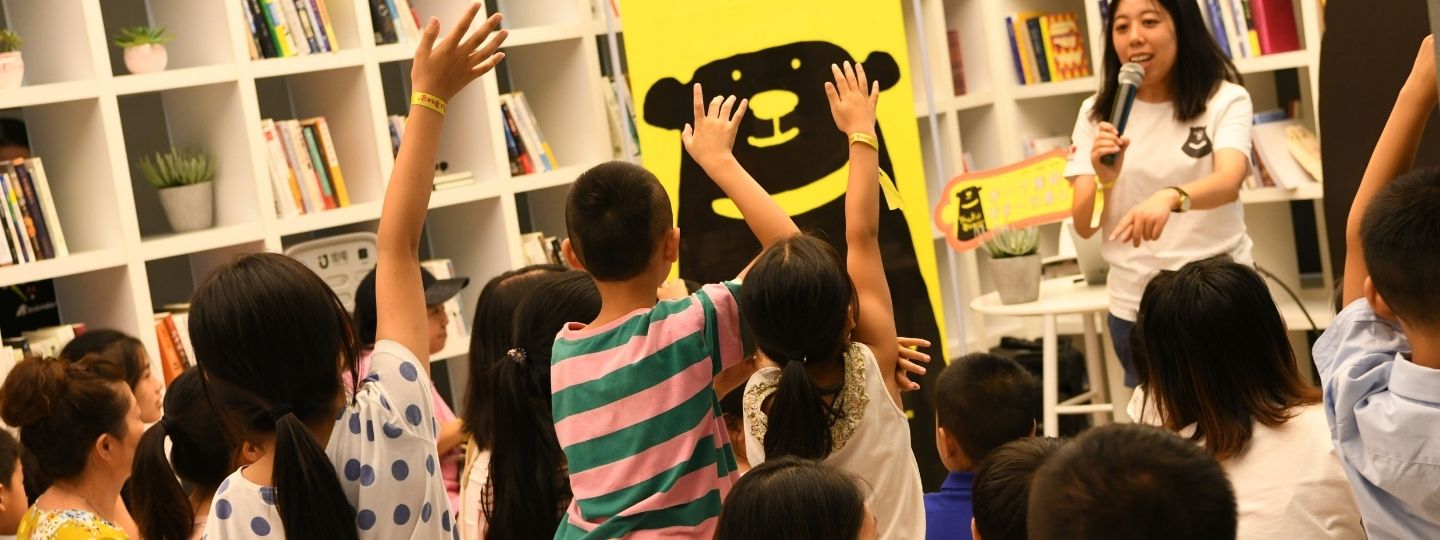An adult with a microphone speaking to a group of primary aged schoolkids – some of who are raising their hands – facing a banner with a cartoon bear on it.