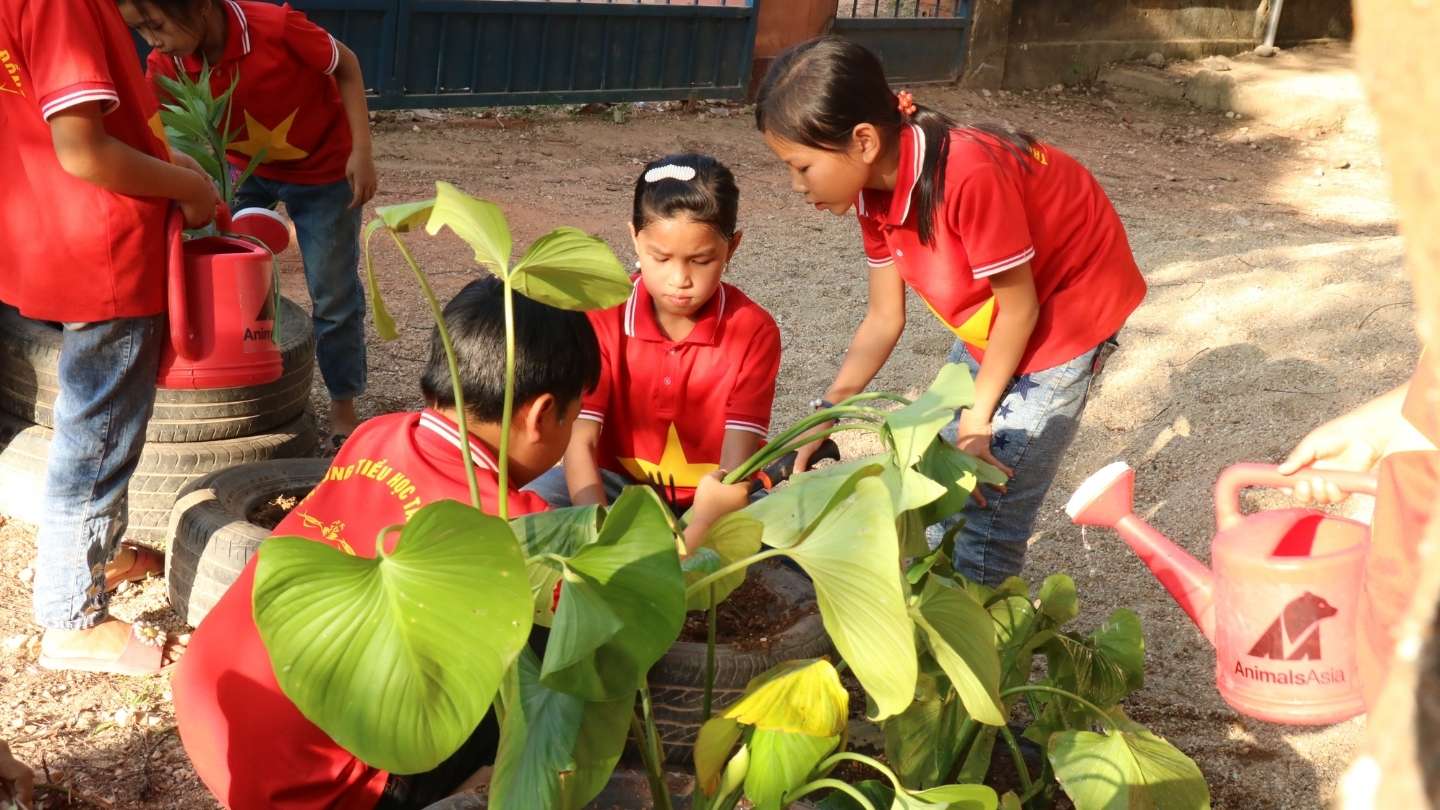 Children in red shirts watering and tending to plants outdoors using red watering cans.