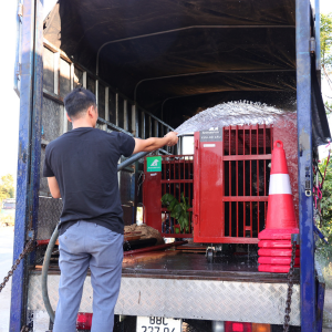 Man in black t-shirt and blue jeans pointing a hose into the back of a small truck, aiming water at red cage with black bear inside.