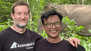 Two men in Animals Asia tshirts stand smiling in a lush forest with an elephant in the background.