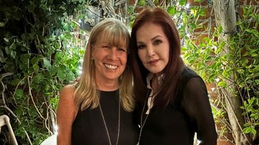 Two women stand closely, smiling and facing the camera.