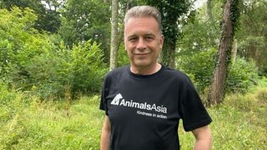 Middle aged man stands in a wood wearing a black t-shirt that says Animals Asia