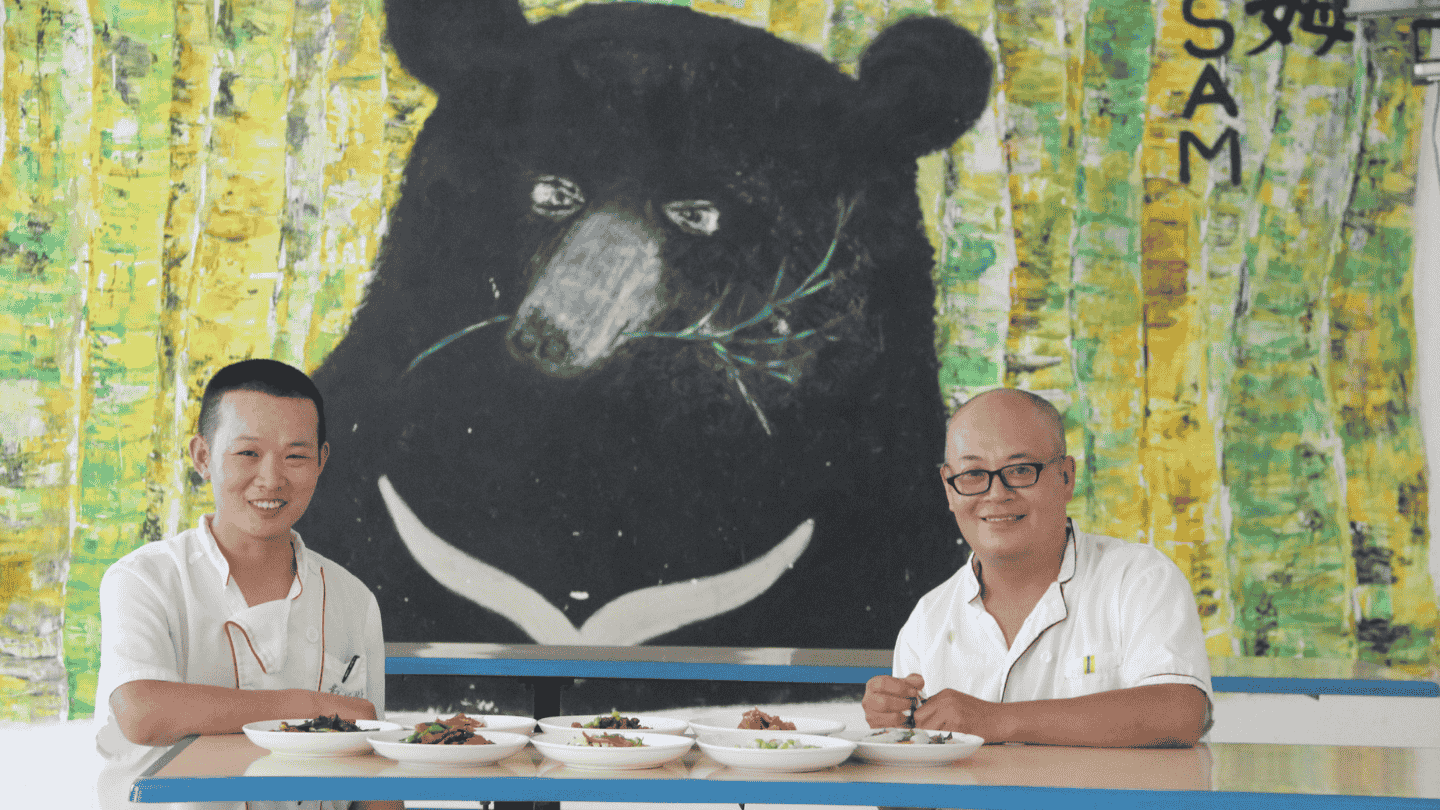 Two chefs sitting behind a tale with dishes of food, looking to camera with mural of Asiatic black bear in the background.