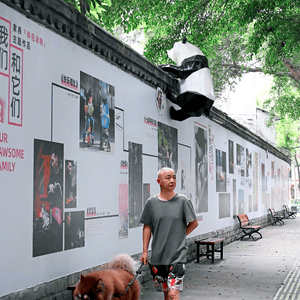Image of man in grey t-shirt and multi-coloured shorts with brown, fluffy dog on a lead waking towards the camera, past high wall covered in images from photographic exhibition