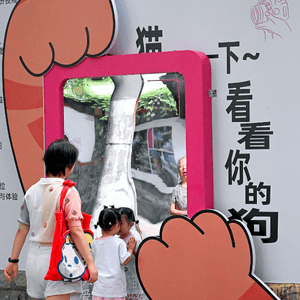 Image of adult woman with short dark hair, and red bag on right shoulder with small girl in pigtails in front of her looking into a red-framed large mirror being held up by orange cat paws.