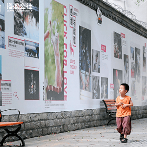 Image of a small boy in orange T-shirt and maroon trousers, sandals walking towards the camera, looking to his right at a high wall covered in images from a photographic exhibition