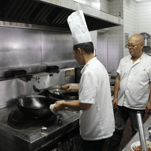Two chefs in steel-appliance kitchen, one wearing tall chef's hat, the other observing him cook