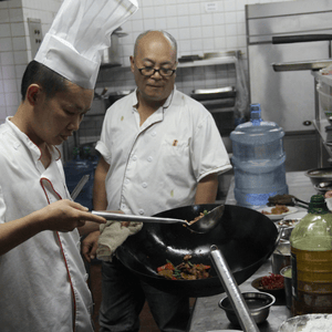 Two chefs in steel-appliance kitchen, one wearing tall chef's hat, the other observing him cook