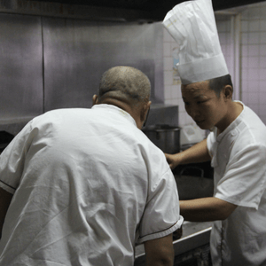 Two chefs in steel-appliance kitchen, one wearing tall chef's hat facing camera, the other with back to camera.
