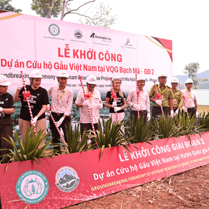 Nine people standing in a row with red and white striped shovels, posing behind plants and in front of a large banner.
