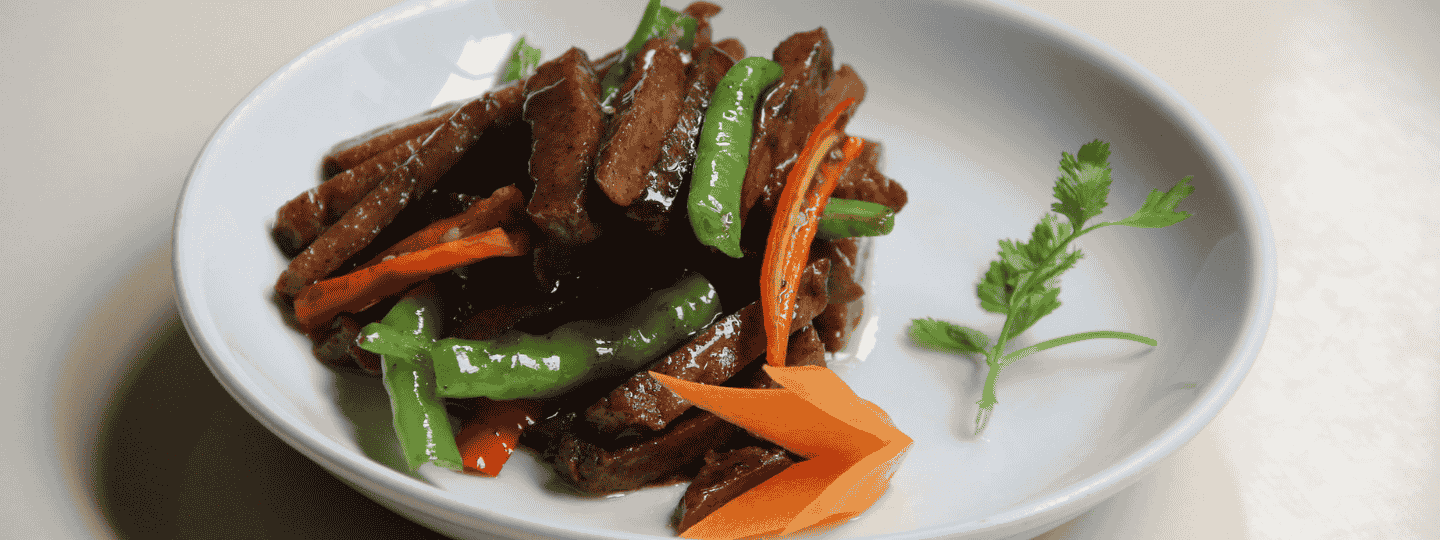 Vegan plant-based meat with red peppers and green beans displayed on a round, white plate.