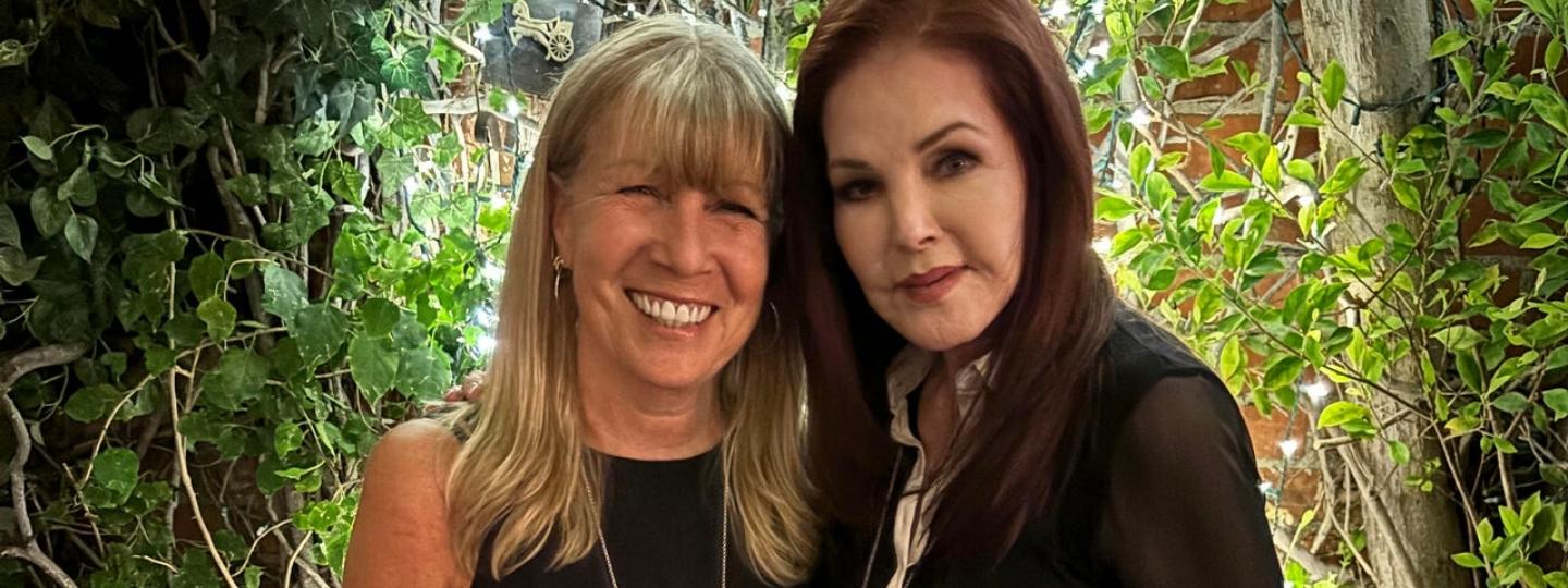 Two women stand closely, smiling and facing the camera.