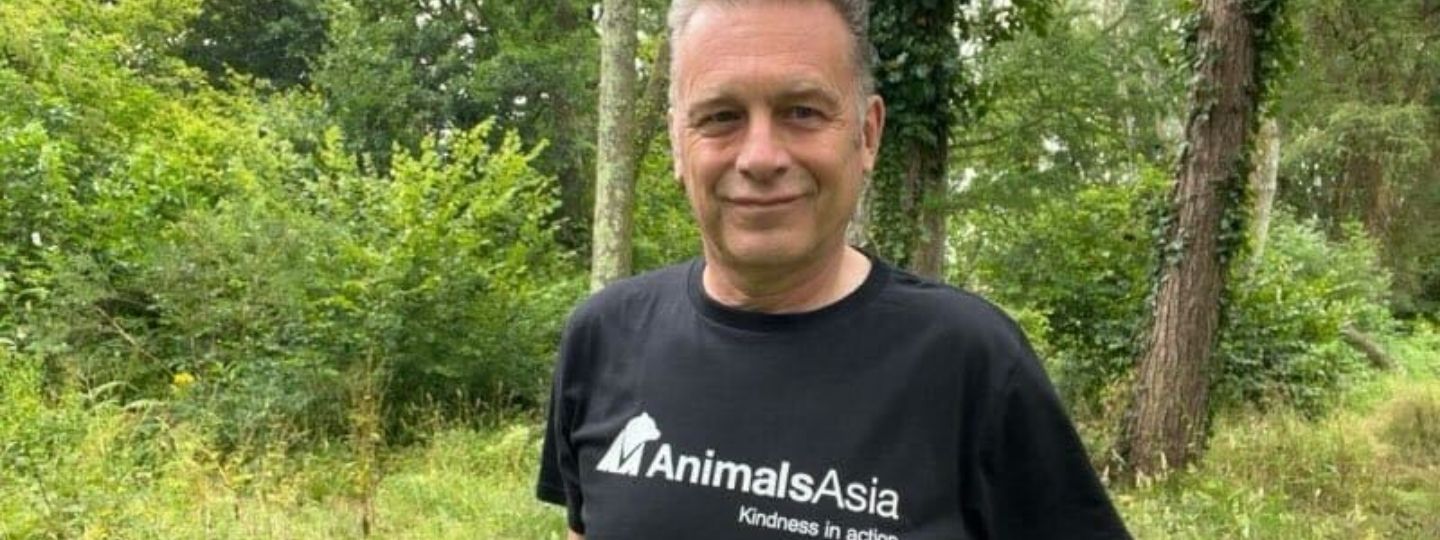 Middle aged man stands in a wood wearing a black t-shirt that says Animals Asia