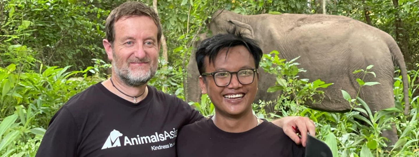 Two men in Animals Asia tshirts stand smiling in a lush forest with an elephant in the background.
