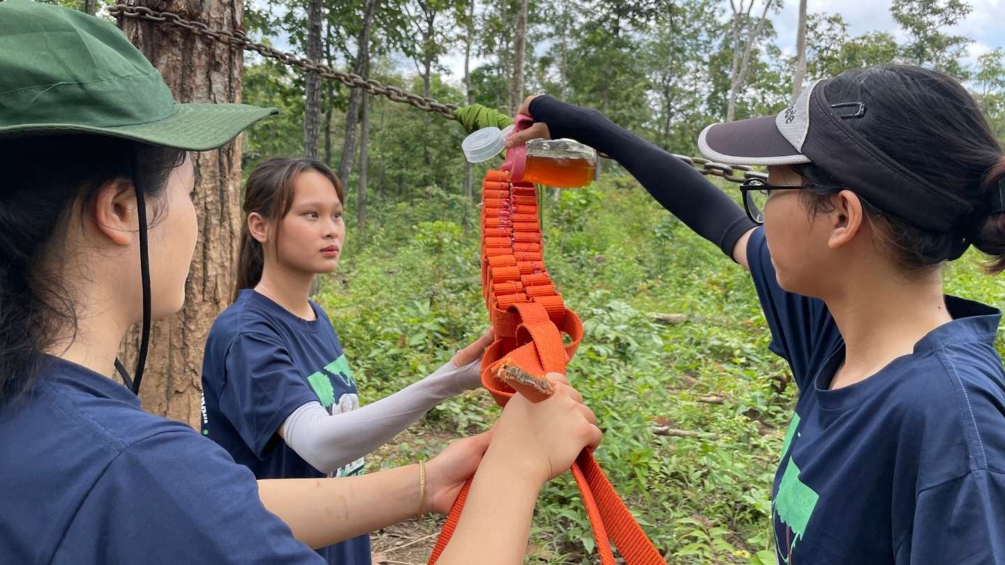 Three women in a lush forest holding an enrichment item as one pours honey onto it